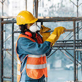 Trabajador de la construcción con casco amarillo y mascarilla manipula una estructura metálica.